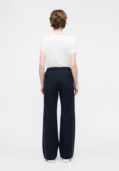 Άτομο με κοντά σγουρά μαλλιά, που φοράει λευκό πουκάμισο με κοντά μανίκια και άνετο μαύρο παντελόνι με λευκές ρίγες, στέκεται με την πλάτη γυρισμένη.