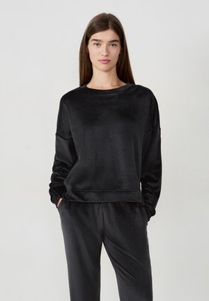 Mujer con cabello largo castaño que lleva una blusa de terciopelo negra de manga larga y pantalones a juego, de pie con las manos en los bolsillos frente a un fondo gris.