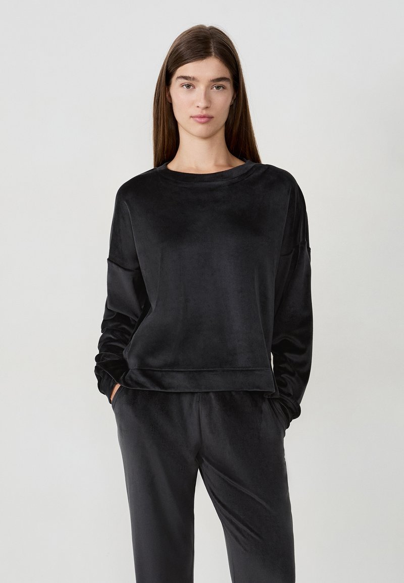Donna con lunghi capelli castani, che indossa un top a maniche lunghe in velluto nero e pantaloni coordinati, in piedi con le mani nelle tasche su uno sfondo grigio.