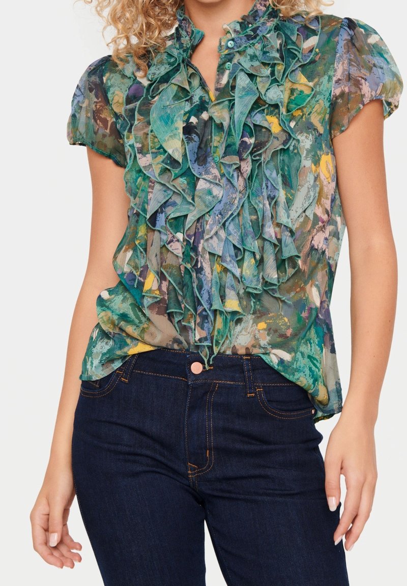 Blouse transparente multicolore avec un devant volants et des manches courtes, dans des teintes de vert, bleu et jaune. Associée à un jean en denim foncé.