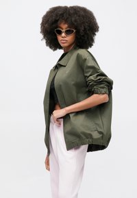 Ελαφρύ μπουφάν σε ελιά πράσινο χρώμα με γιακά, ελαστικά μανίκια και πλαϊνές τσέπες, φορεμένο πάνω από μαύρο crop top και ροζ παντελόνι.