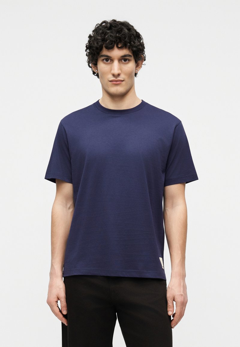 Νεαρός με σγουρά μαλλιά που φοράει μια μονόχρωμη μπλε ναυτική κοντομάνικη μπλούζα και μαύρο παντελόνι, στέκεται μπροστά σε λευκό φόντο.