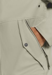 Lichtgrijze jacket met een ritszak, textuurstof en een zwarte drukknoopdetail, met een close-up van de hand in de zak.