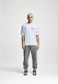 Tricou alb cu mânecă scurtă și logo pe piept, asortat cu pantaloni de trening gri și adidași negri cu accente albe.