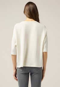 Haut blanc, ample, à manches courtes et col rond, avec une texture lisse et des fentes sur les côtés, associé à un jean gris ajusté.