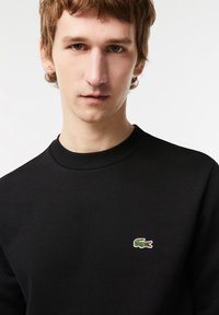 Sudadera negra con cuello redondo, que presenta un pequeño logo de cocodrilo bordado en verde y rojo en el lado izquierdo del pecho. Textura suave, diseño casual.