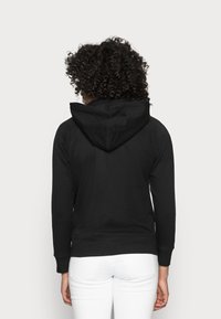 En person med lockigt hår står vänd bort med en svart hoodie och vita byxor mot en enkel ljus bakgrund.