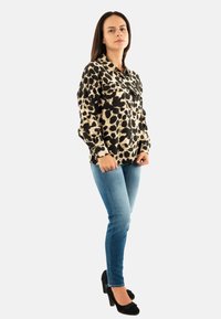 Blusa a macchie nera e beige, maniche lunghe, con bottoni frontalmente, abbinata a jeans blu e scarpe con tacco nere. Sfondo bianco uniforme.