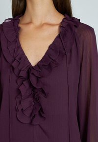 Blusa viola con scollatura a V profonda, ornata da dettagli arricciati lungo il collo e bottoni, realizzata in un tessuto leggero.
