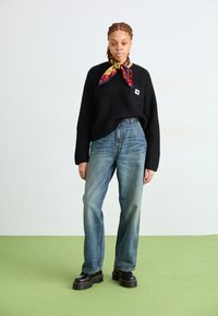 Maglione nero a costine con tasca frontale, jeans blu a gamba dritta, sciarpa a fantasia gialla e rossa, stivali neri chunky, ambiente interno.