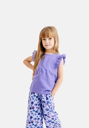 Junges Mädchen mit langen blonden Haaren, das ein lila Oberteil mit Volantärmeln und lockere Hosen mit Blumenmuster trägt, die Hände in den Taschen.