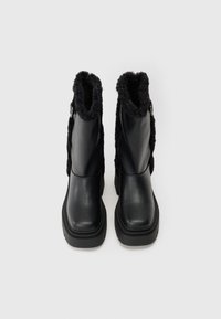 Botas de tornozelo em pele sintética preta com um punho em pelúcia sintética preta texturizada, bico quadrado e sola de borracha espessa. Apresenta hardware decorativo nas laterais.