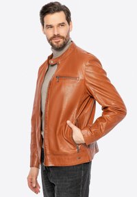 Giacca di pelle marrone con una texture liscia, tasca con zip sul petto, tasche laterali e colletto con chiusura a clip. Indossata sopra un maglione grigio.