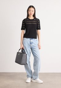 Black knit top with cut-out pattern, paired with light blue baggy jeans and a small, patterned, black handbag. White sneakers complete the outfit.