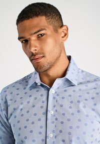 Camicia button-up blu chiaro con un sottile motivo a pois in diverse sfumature di blu, dotata di un colletto classico e una texture liscia.