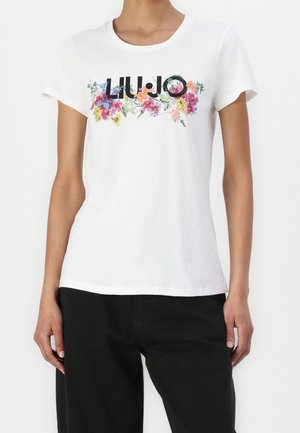 Mujer con una camiseta blanca de manga corta con el texto "LIU·JO" y un colorido diseño floral, combinada con pantalones negros.