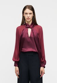 Γυναίκα φοράει μπορντό σατέν μπλούζα με κόμπο στο λαιμό και διαφανή μακριά μανίκια, συνδυασμένη με μαύρο παντελόνι με φαρδιά μπατζάκια και ψηλή μέση.