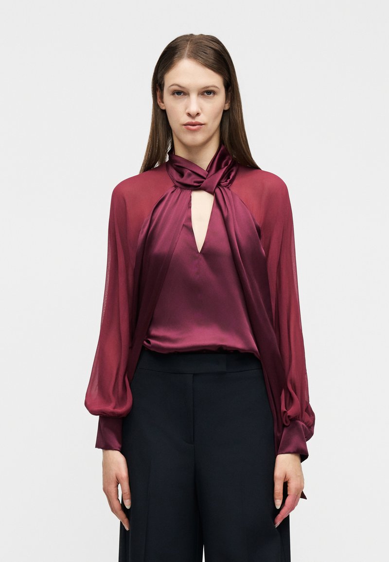 Γυναίκα φοράει  μπορντό σατέν μπλούζα με κόμπο στο λαιμό και διαφανή μακριά μανίκια, συνδυασμένη με μαύρο παντελόνι με φαρδιά μπατζάκια και ψηλή μέση.