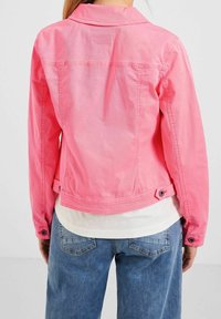 Person wearing a bright pink denim jacket over a white shirt and blue jeans, shown from the back against a plain background.