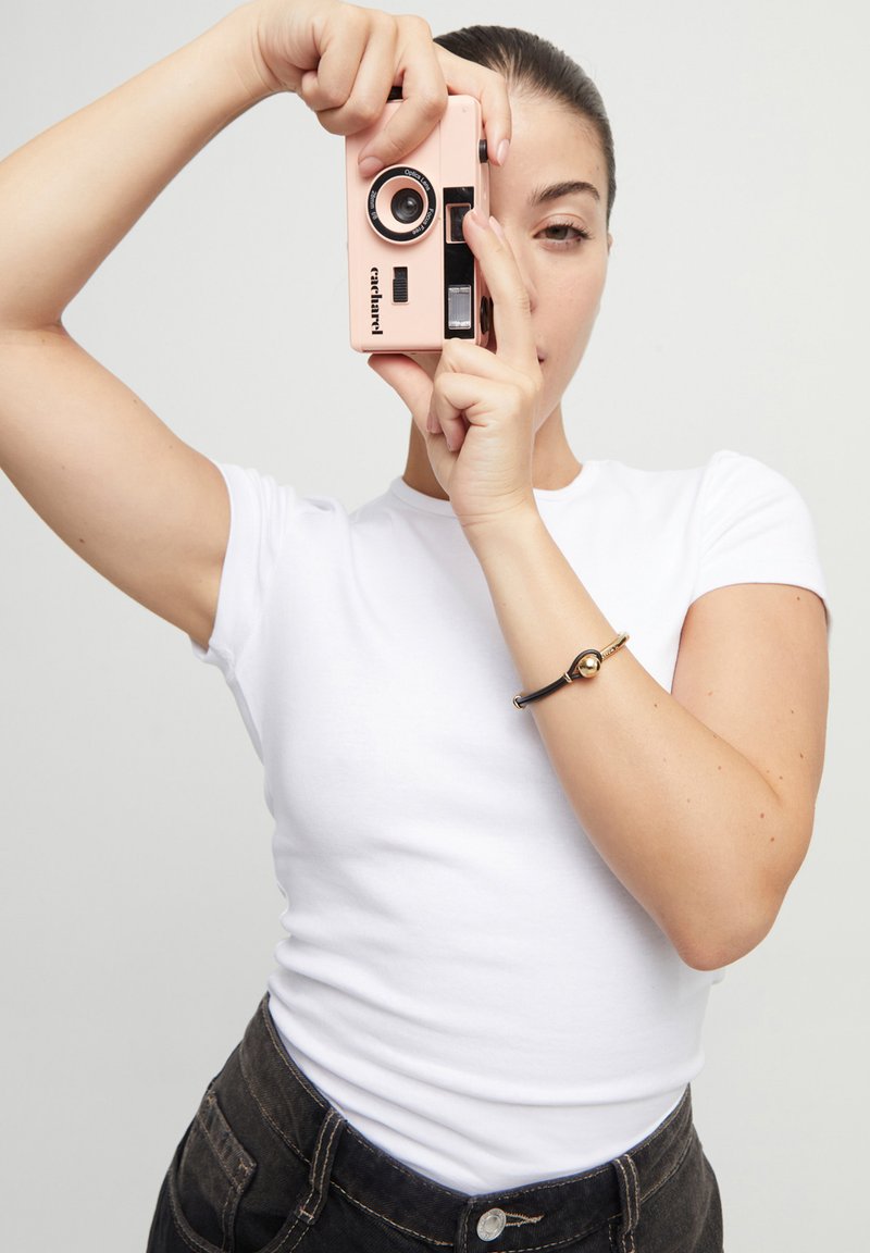 Cámara instantánea rosa con acentos negros sostenida por una persona que lleva una camiseta blanca. La pulsera presenta una esfera dorada sobre una banda negra.