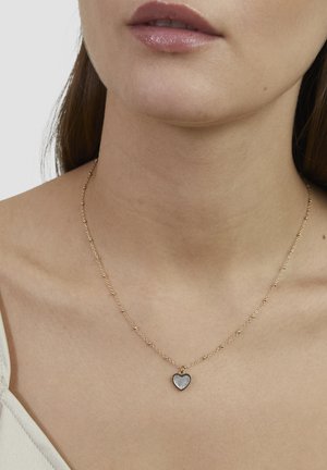Goldene Halskette mit einem herzförmigen Anhänger, der ein grau-weißes Zentrum hat, und auf dem Schlüsselbein liegt.