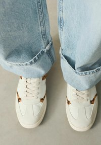 White sneaker with light brown animal print accents, textured leather material, and beige laces, paired with light blue, rolled denim jeans.