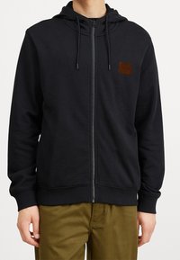 Felpa nera con zip realizzata in tessuto morbido, con cappuccio, tasche frontali e un'etichetta con logo marrone sul lato sinistro del petto. Texture liscia, vestibilità comoda.