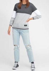 Felpa grigia con maniche a contrasto bianche e grigie chiare, polsini e orlo a costine. Abbinata a jeans azzurri chiari strappati e scarpe da ginnastica nere.