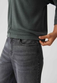 Maglione a coste verde scuro con orlo arrotondato, indossato sopra jeans in denim grigi. La mano regola il bordo inferiore del maglione, mostrando lunghezza e vestibilità.