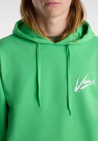 Osoba z długimi włosami ubrana w jasną zieloną bluzę Vans z kapturem, z troczkami i białym logo Vans na piersi.