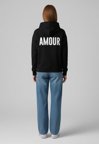 Felpa nera con cappuccio con la scritta "AMOUR" stampata in bianco sul retro, abbinata a jeans a gamba larga blu chiaro e sneakers bianche.