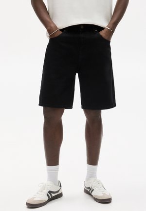 Personne portant un short noir au genou, des chaussettes blanches et des baskets blanches avec des détails noirs, debout les mains dans les poches.