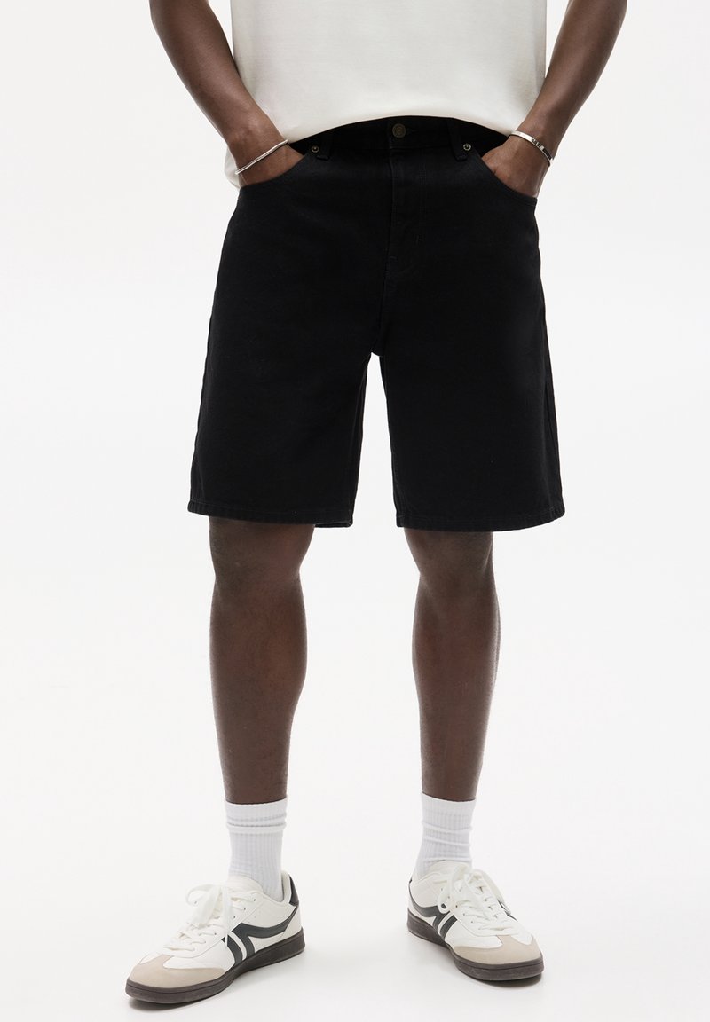 Personne portant un short noir au genou, des chaussettes blanches et des baskets blanches avec des détails noirs, debout les mains dans les poches.