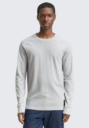 Junger Mann trägt ein hellgraues strukturiertes Langarmshirt und dunkelblaue Jeans, steht frontal vor schlichtem Hintergrund.