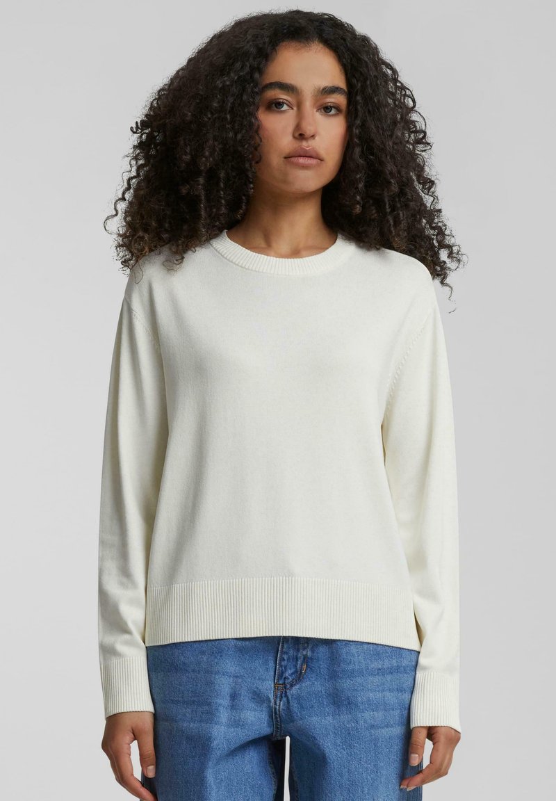 Pull léger beige à manches longues, avec des poignets et un ourlet côtelés. Design simple avec un col rond. Porté avec un jean en denim bleu. Texture douce.
