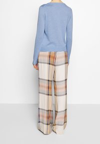 Person in einem hellblauen Langarmtop und weit geschnittenen Karohosen in Beige, Orange und Schwarz, der vor einem schlichten weißen Hintergrund steht.
