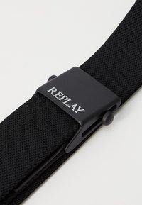 Cintura in tessuto intrecciato nero con fibbia in plastica nera opaca con la scritta "REPLAY" in argento. La fibbia ha un design semplice e piatto.