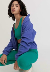 Paarse fleece zipjack heeft een groene sporttop en bijpassende groene legging eronder. Het model gaat in squat houding, met lichtgekleurde sneakers aan.