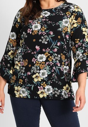 Frau trägt eine schwarze Bluse mit weißen, gelben und rosa Blumenmustern und Dreiviertelärmeln, kombiniert mit dunkelblauer Hose.