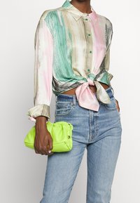 Monivärinen, raidallinen silkkipaita, jossa on solmittu etuosa, sisältäen vihreitä, vaaleanpunaisia ja beigen sävyjä. Käsi, joka kantaa kirjavaa, kirkkaan vihreää käsilaukkua.