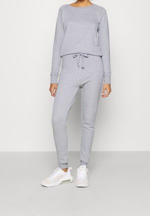 Žena oblečená ve světlé šedé mikině s dlouhými rukávy a odpovídajících teplácích, s bílými sportovními botami, stojící před jednobarevným pozadím.