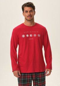 Czerwona koszula z długim rękawem z białymi nadrukami płatków śniegu oraz napisem "ŚWIĘTA W TRAKCIE ŁADOWANIA". Połączona z spodniami w kratkę w zielono-czerwonym kolorze.