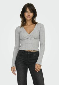 Grijze geribbelde lange mouwen wikkeltop met een V-hals en een scalloped zoom, gecombineerd met zwarte high-waisted jeans, weergegeven op een neutrale achtergrond.