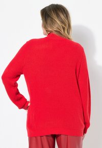 Pull en tricot rouge avec une texture côtelée, manches longues et col haut. Le dos présente une coupe décontractée et de légents fentes sur les côtés au niveau de l'ourlet.