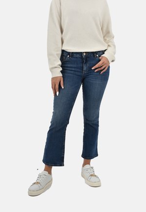 Jeans in denim blu scuro con orlo svasato, dotati di passanti per cintura e un leggero motivo di scolorimento. Abbinati a sneaker bianche e a un maglione leggero.