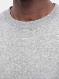 Close-up af en persons hals og den øverste del af en lysegrå crewneck sweater med ribstrikket krave.