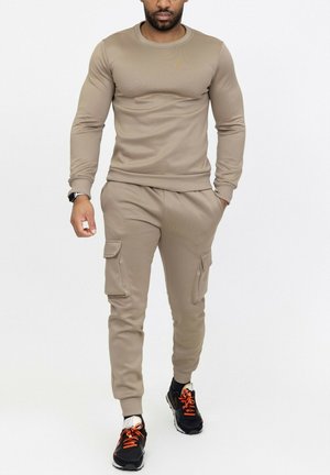 Homme portant un sweat-shirt beige à manches longues et un pantalon cargo avec des baskets noires aux lacets orange, marchant devant un fond blanc uni.
