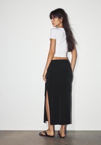 Black maxi skirt with side slit, paired with a fitted white t-shirt. Model stands against a plain background, wearing flat sandals.