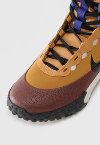 Barna és bézs Nike sneaker texturált orrrésszel, fekete talppal, fekete és lila cipőfűzőkkel, valamint részleges fekete Nike logóval fehér háttérrel.