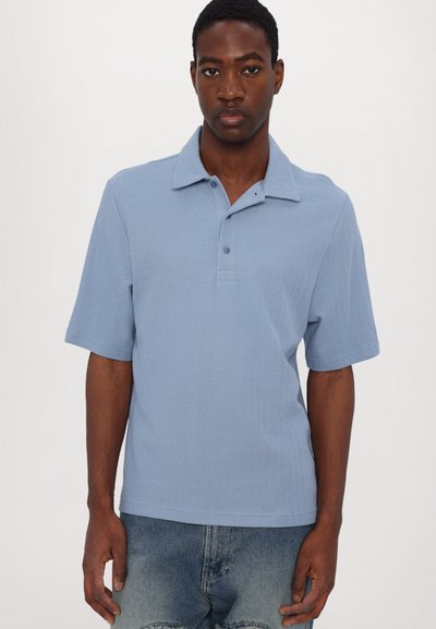 Šviesiai mėlynas trumpomis rankovėmis polo marškinėliai su apykakle, trijų mygtukų užsegimu ir tekstūruota medžiaga. Dėvimi virš išblukusių džinsinių šortų.